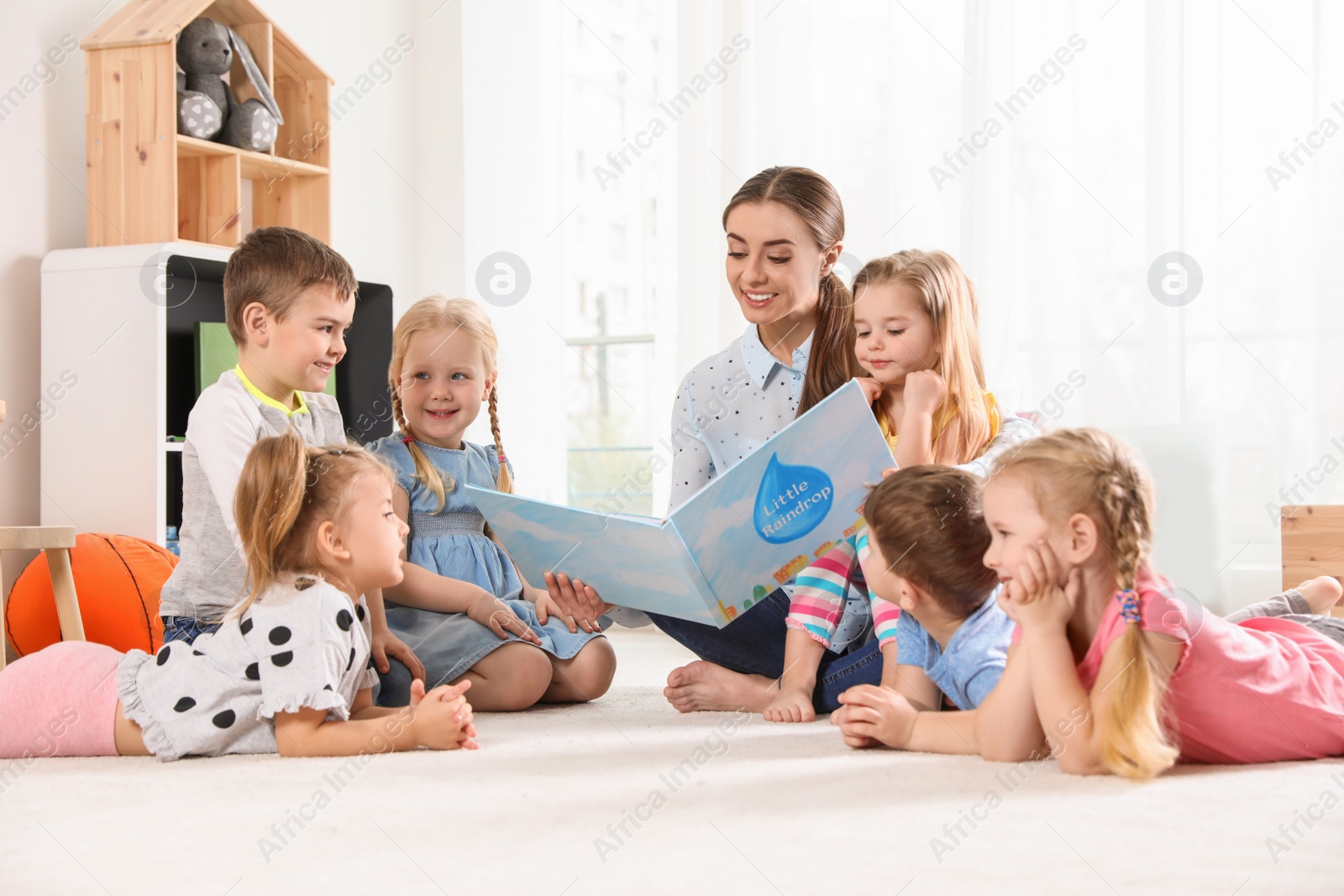 Photo of Kindergarten teacher reading book to children indoors. Learning and playing