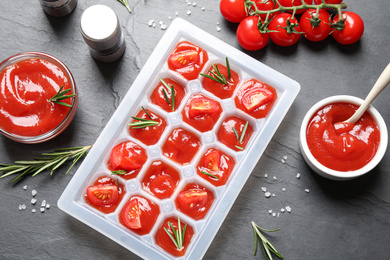 Ice cube tray with tomatoes, sauce and fresh rosemary on grey table, flat lay