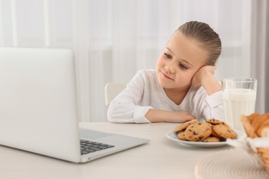 Photo of Little girl using laptop while having breakfast at table indoors. Internet addiction