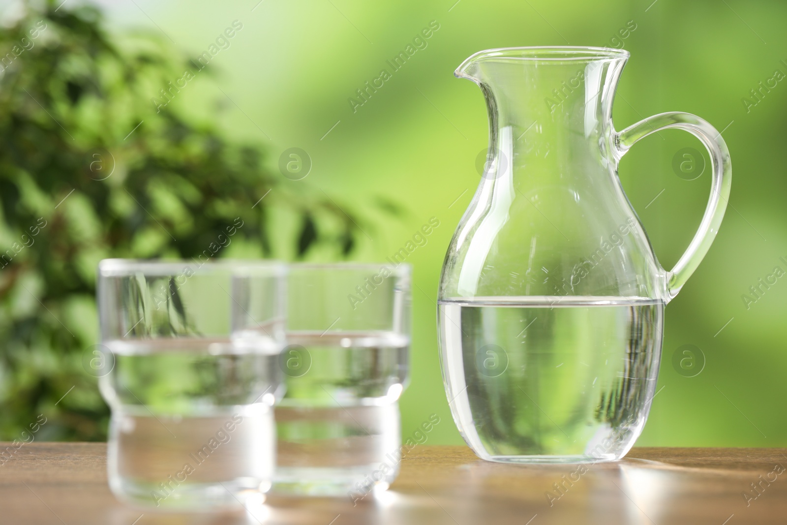 Photo of Jug and glasses with clear water on wooden table against blurred green background, closeup