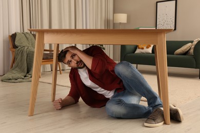 Photo of Scared man hiding under table in living room during earthquake