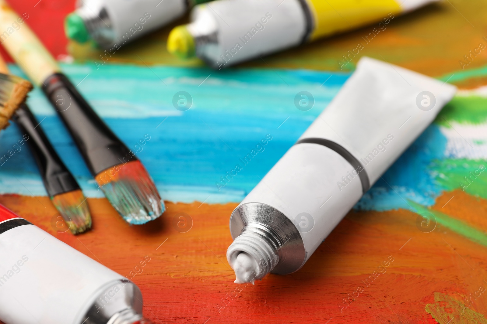 Photo of Tubes of colorful oil paints and brushes on canvas with abstract painting, closeup