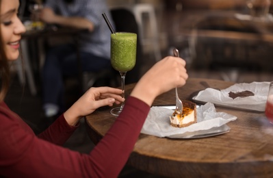 Pretty young woman with cocktail and cake at table in cafe, view from outdoors through window