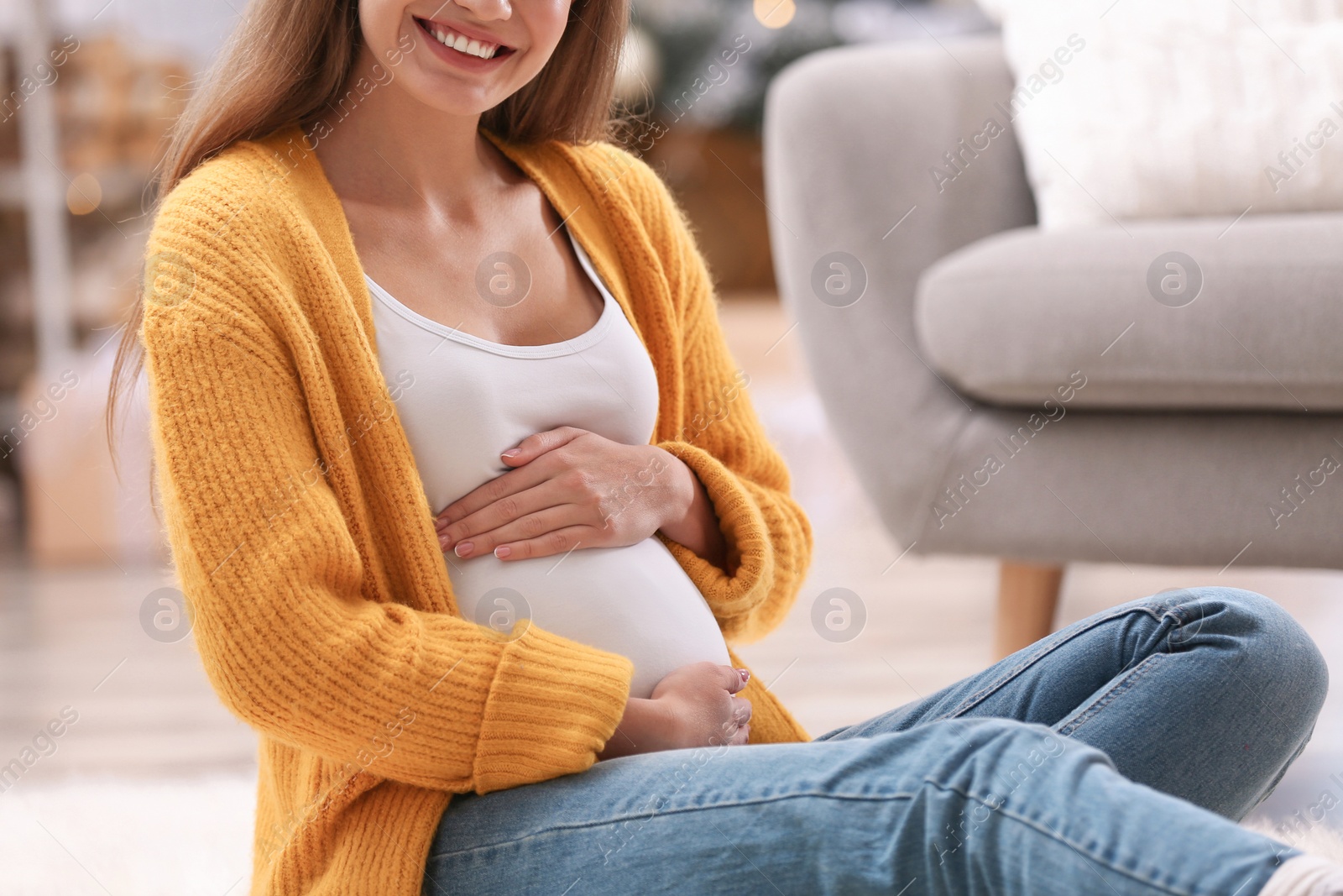 Photo of Young pregnant woman sitting on floor in room decorated for Christmas, closeup