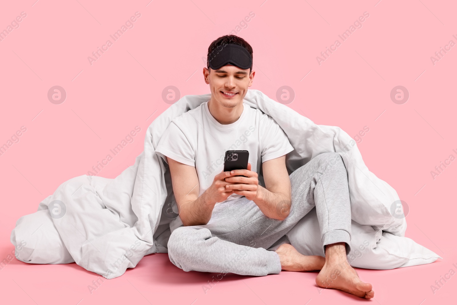 Photo of Happy man in pyjama with sleep mask, blanket and smartphone on pink background