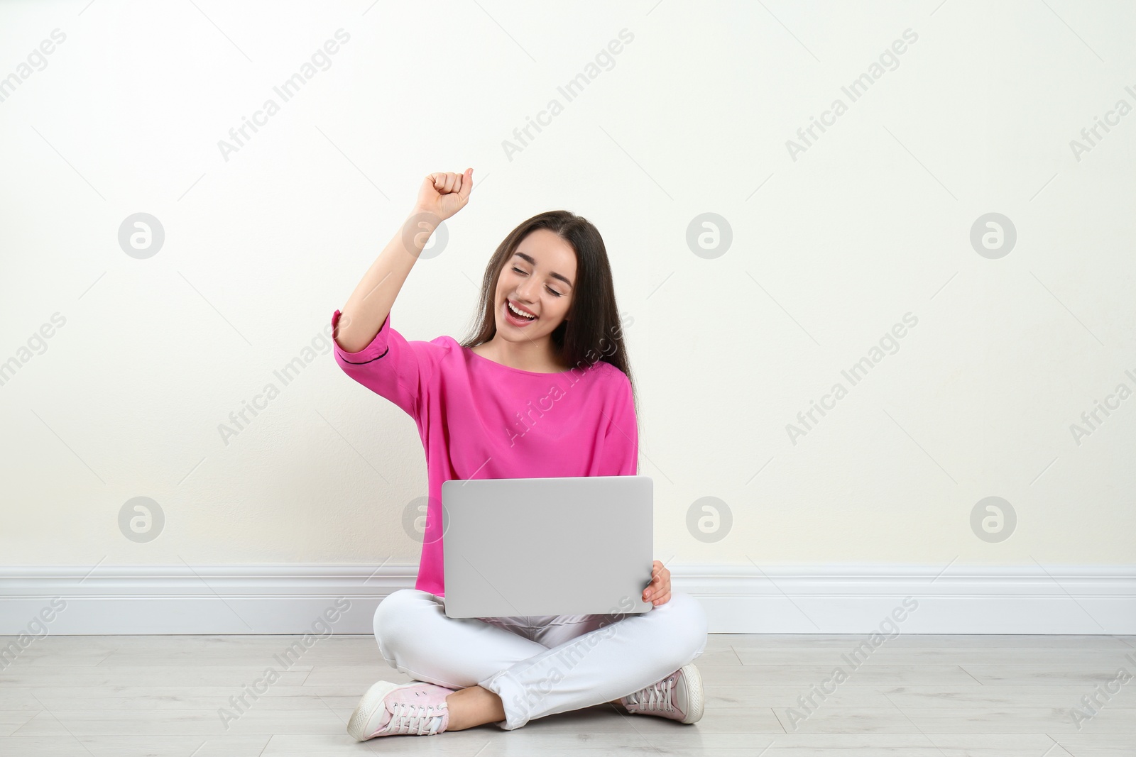Photo of Happy young woman with laptop sitting on floor near light wall indoors