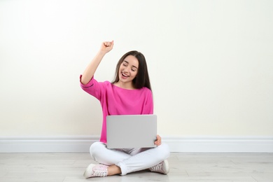 Happy young woman with laptop sitting on floor near light wall indoors