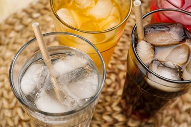 Glasses of different refreshing soda water with ice cubes and straws on wicker mat, closeup