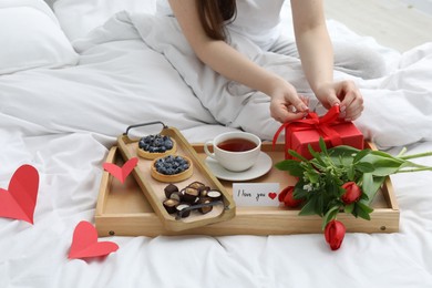 Photo of Tasty breakfast served in bed. Woman with gift box, tea, desserts, flowers and I Love You card at home, closeup