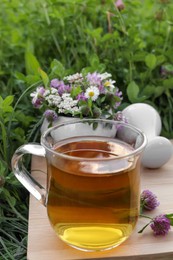 Photo of Cup of aromatic herbal tea, pestle and ceramic mortar with different wildflowers on wooden board in meadow
