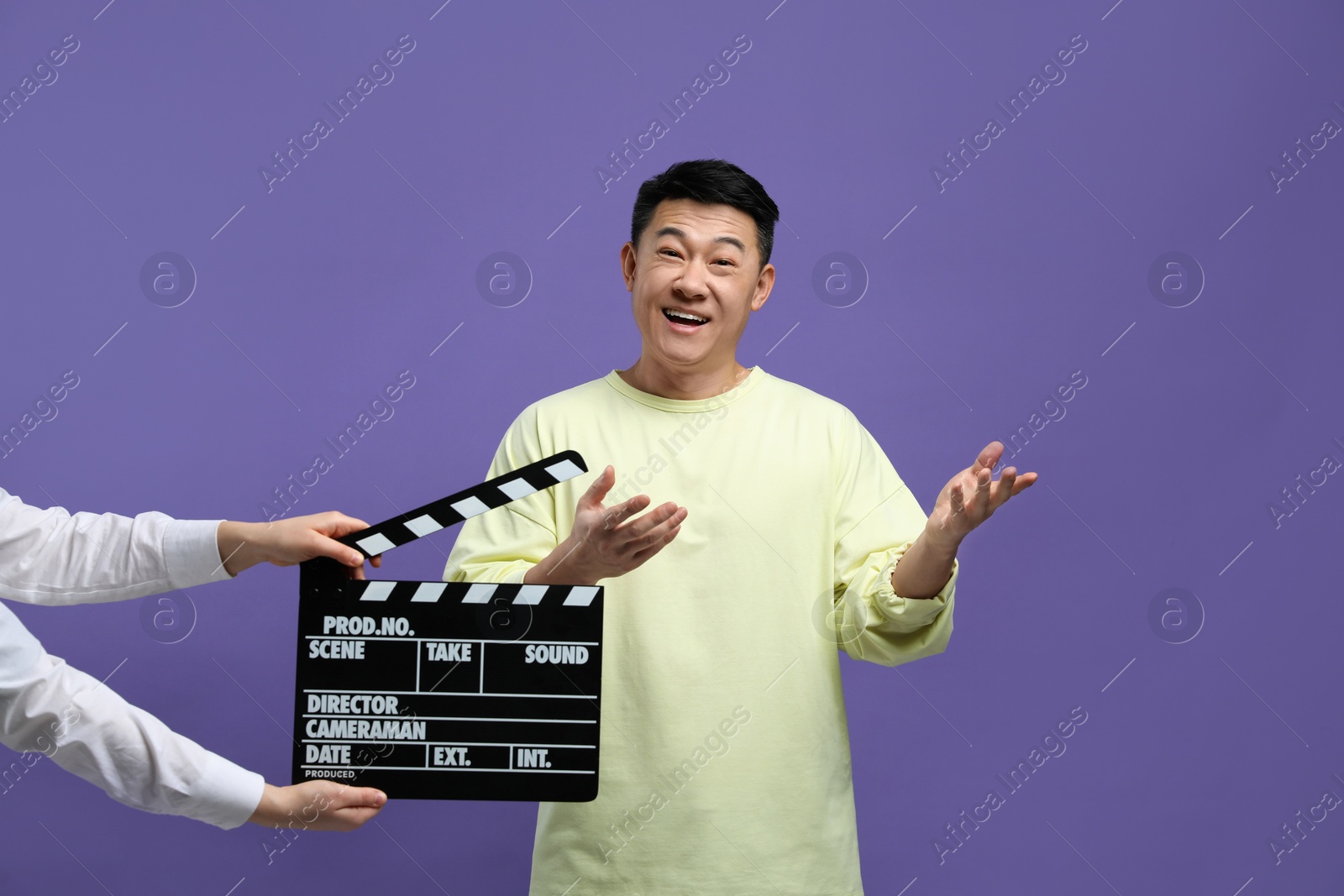 Photo of Happy asian actor performing while second assistant camera holding clapperboard on purple background. Film industry