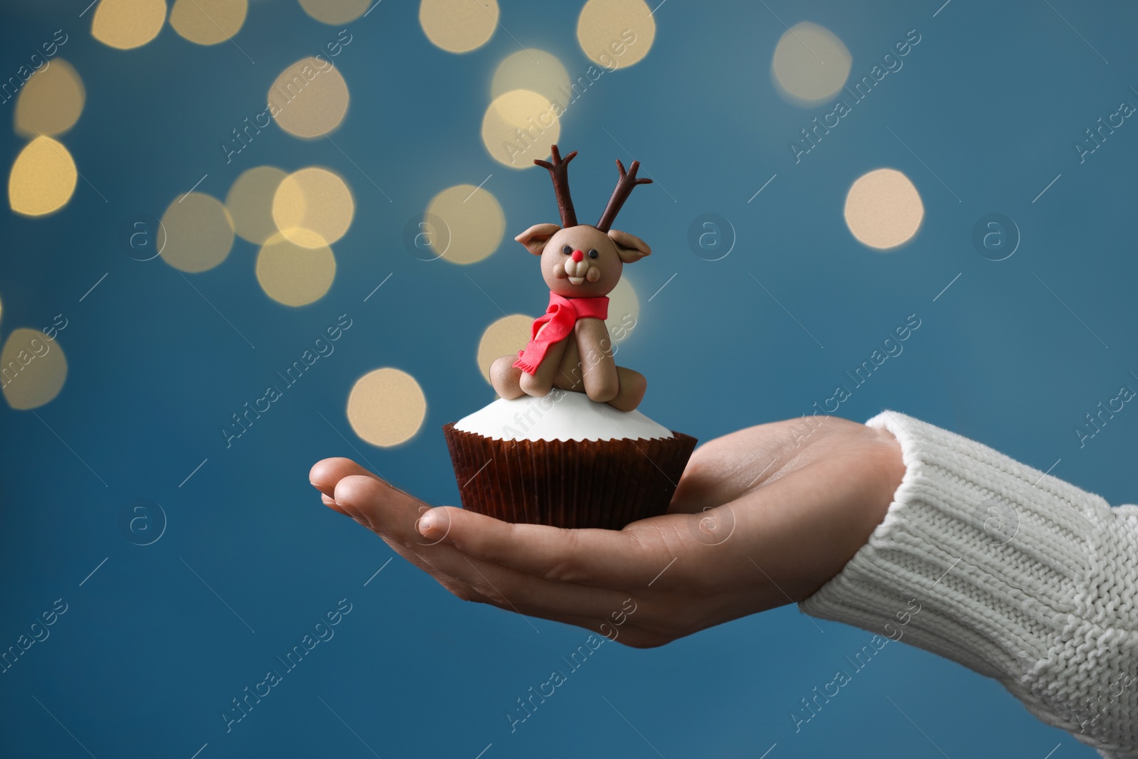 Photo of Woman holding tasty Christmas cupcake with reindeer figure against blurred festive lights, closeup