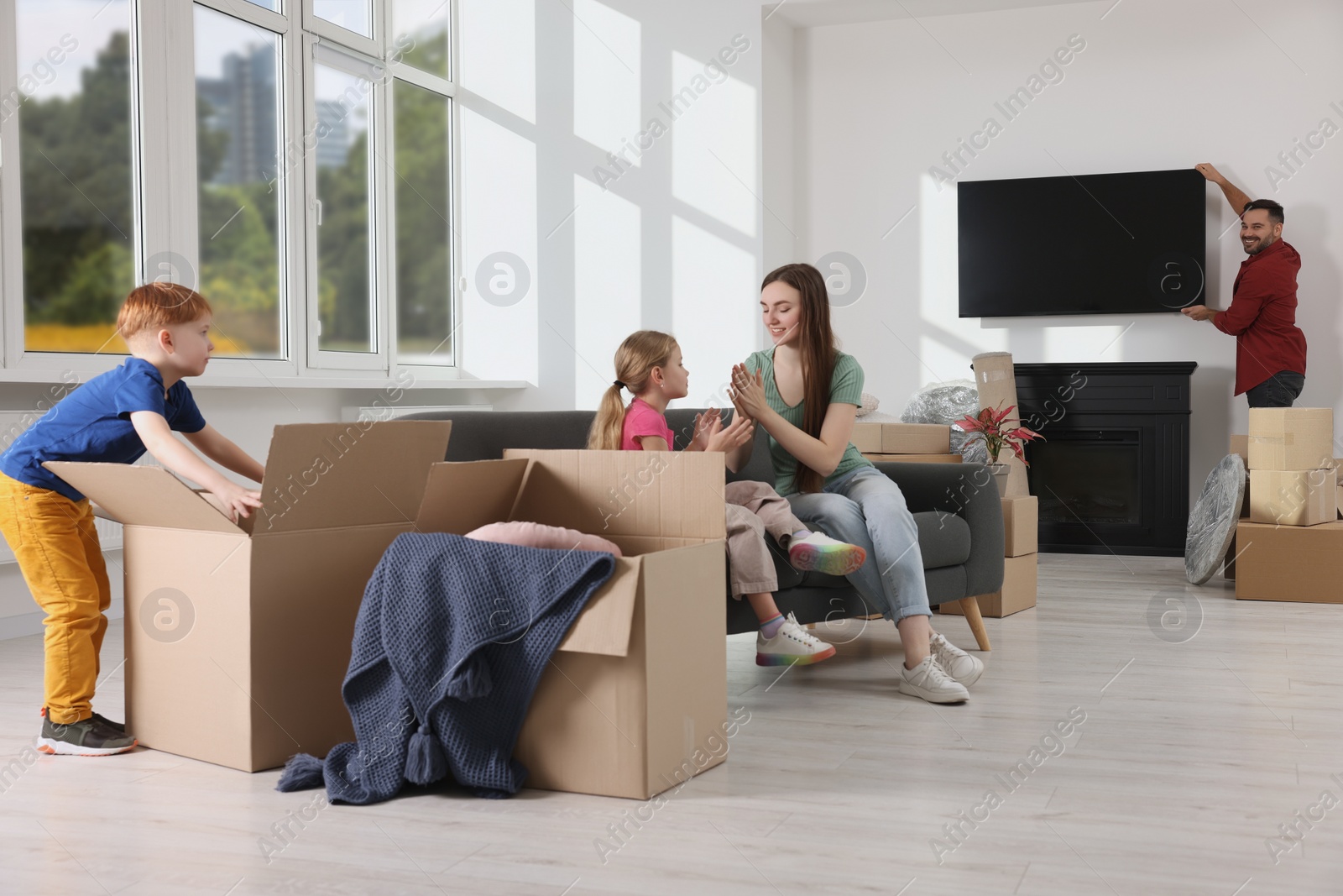 Photo of Family settling into new home. Dad setting up TV, happy mom and daughter playing on couch, son unpacking his boxes. Moving day