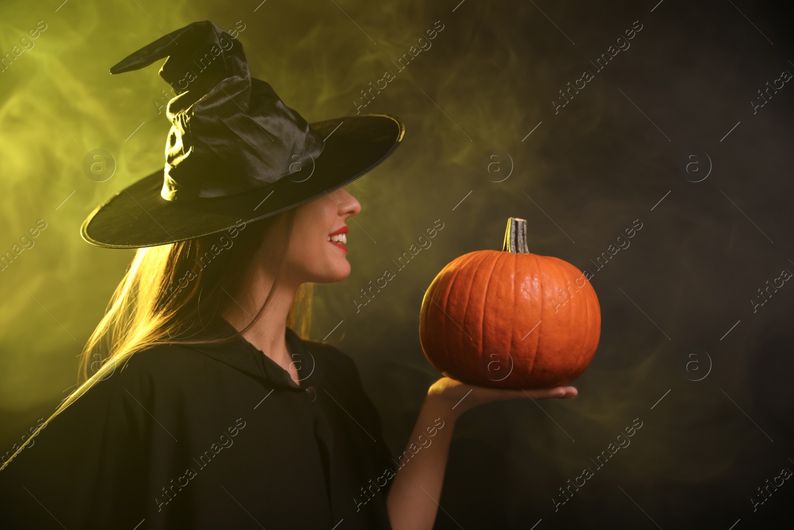 Photo of Young woman wearing witch costume with pumpkin in smoke cloud on dark background. Halloween party