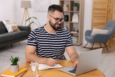 Young man writing in notebook while working on laptop at wooden table indoors