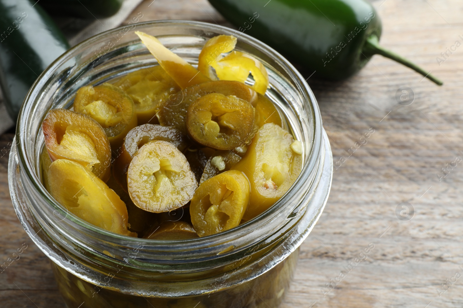 Photo of Glass jar with slices of pickled green jalapeno peppers on wooden table, closeup