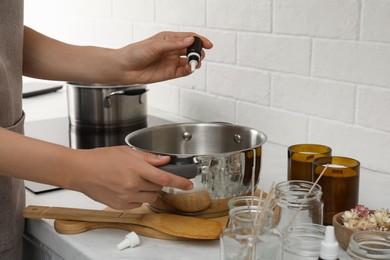 Woman adding essential oil into pot with melted wax at kitchen counter, closeup. Making homemade candles