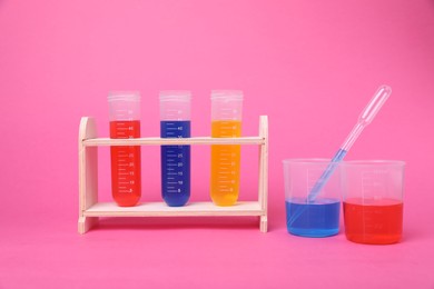 Photo of Beakers and test tubes with colorful liquids in wooden stand on bright pink background. Kids chemical experiment set
