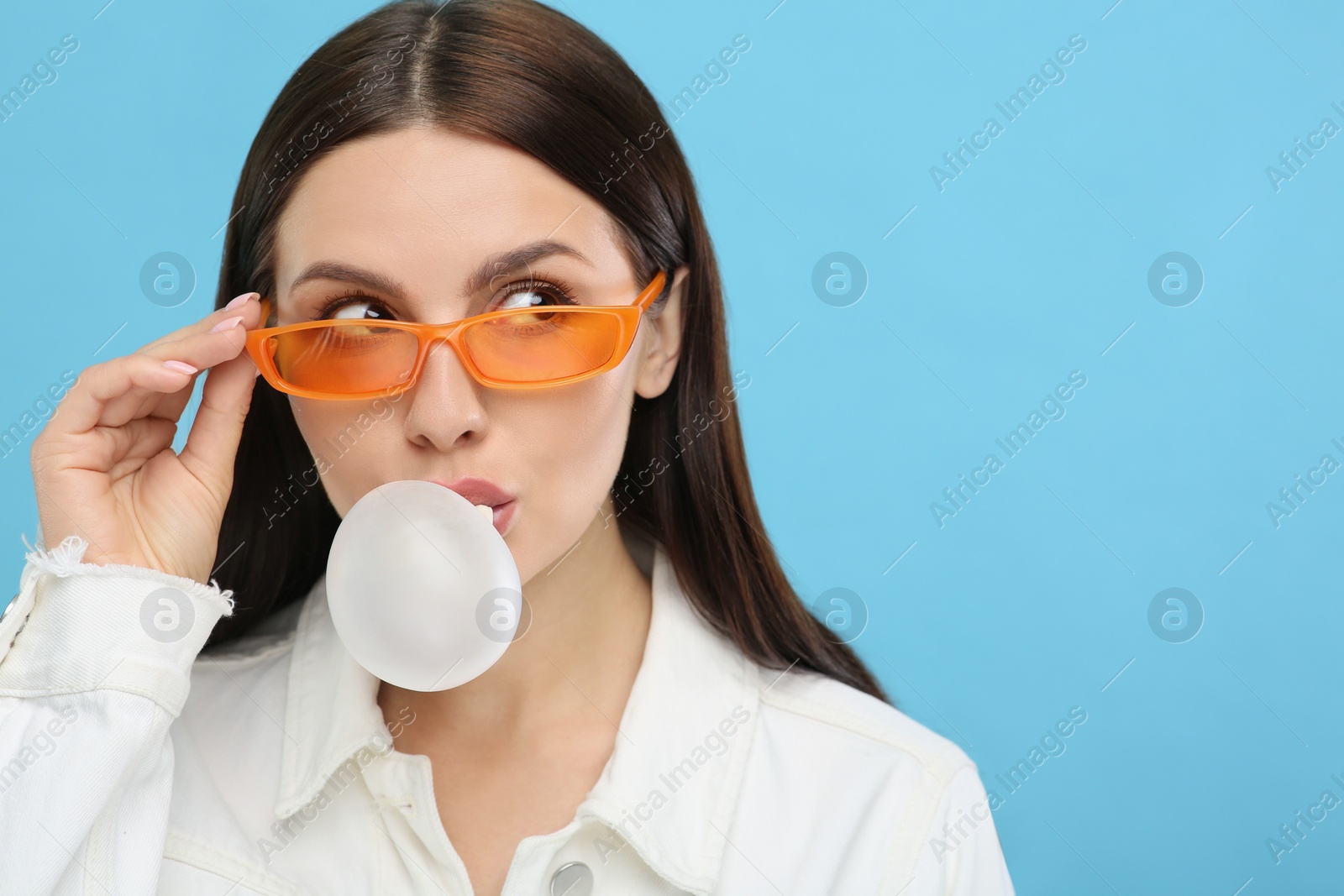 Photo of Beautiful woman in sunglasses blowing bubble gum on light blue background, space for text