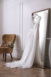 Photo of Elegant wedding dress hanging on large mirror in room