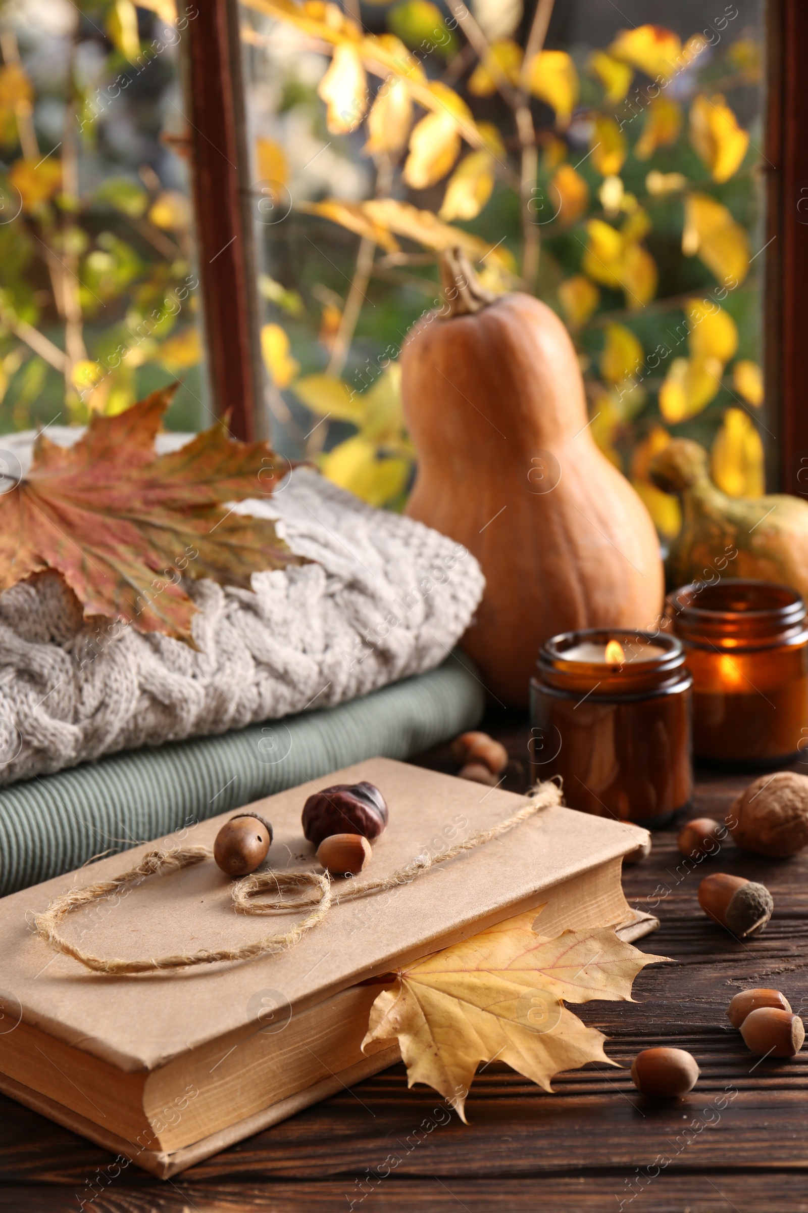 Photo of Book with autumn leaf as bookmark, acorns, scented candle and warm sweaters on wooden table near window, space for text