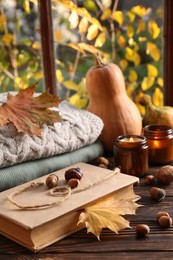 Book with autumn leaf as bookmark, acorns, scented candle and warm sweaters on wooden table near window, space for text