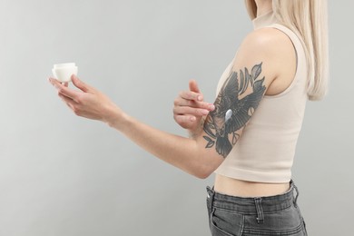 Tattooed woman applying cream onto her arm on gray background, closeup