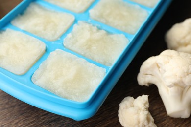 Cauliflower puree in ice cube tray and fresh cauliflower on wooden table, closeup