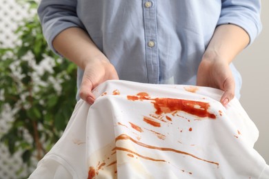 Photo of Woman holding shirt with stain of sauce on blurred background, closeup