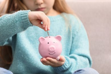 Photo of Cute little girl putting money into piggy bank indoors, closeup