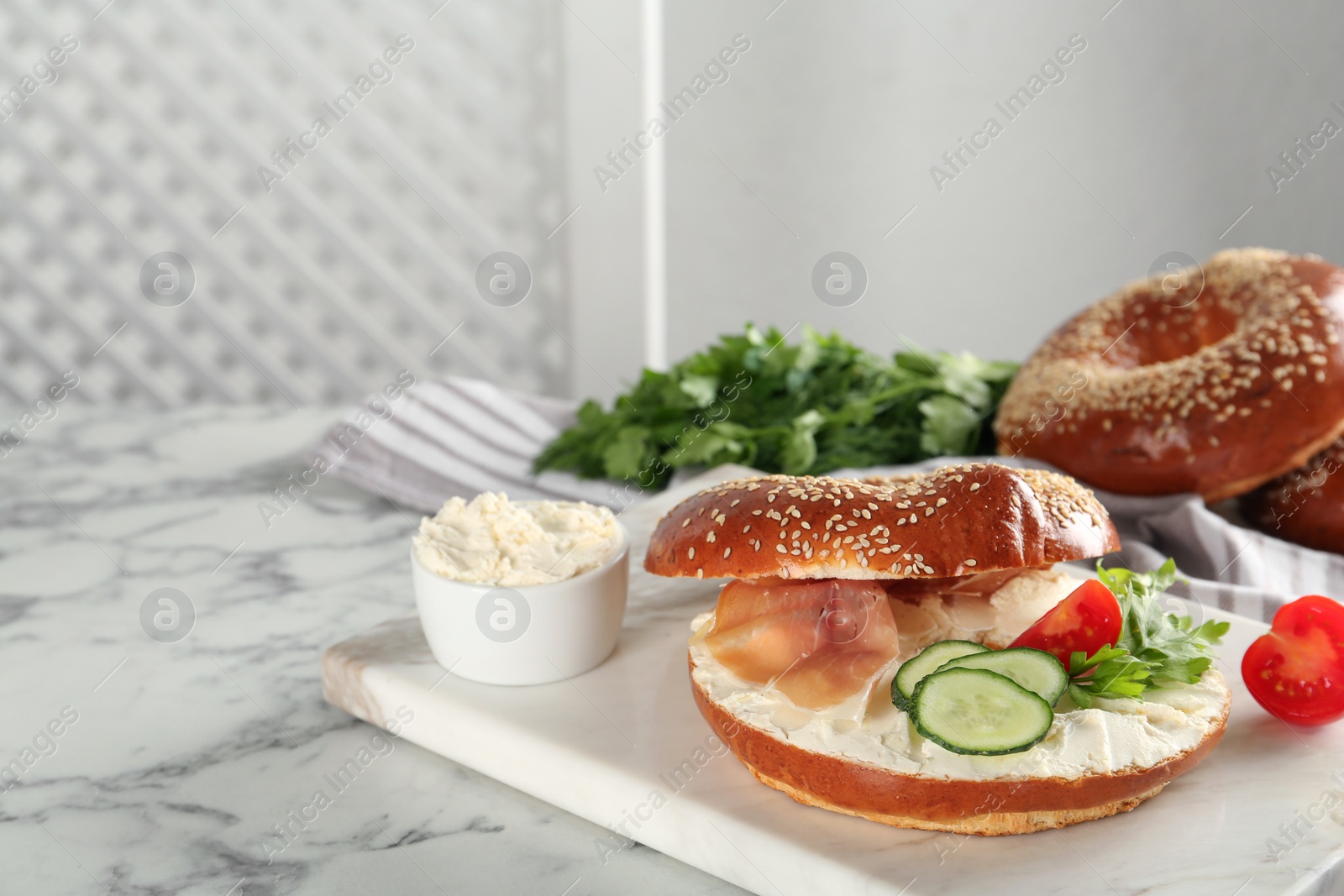 Photo of Delicious bagel with cream cheese, jamon, cucumber, tomato and parsley on white marble table. Space for text