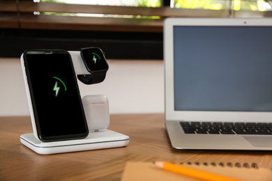 Mobile phone, earphones and smartwatch charging with wireless pad on wooden desk. Modern workplace device