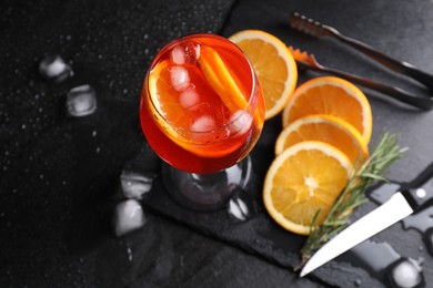 Photo of Glass of tasty Aperol spritz cocktail with orange slices, ice cubes and rosemary on black table, above view