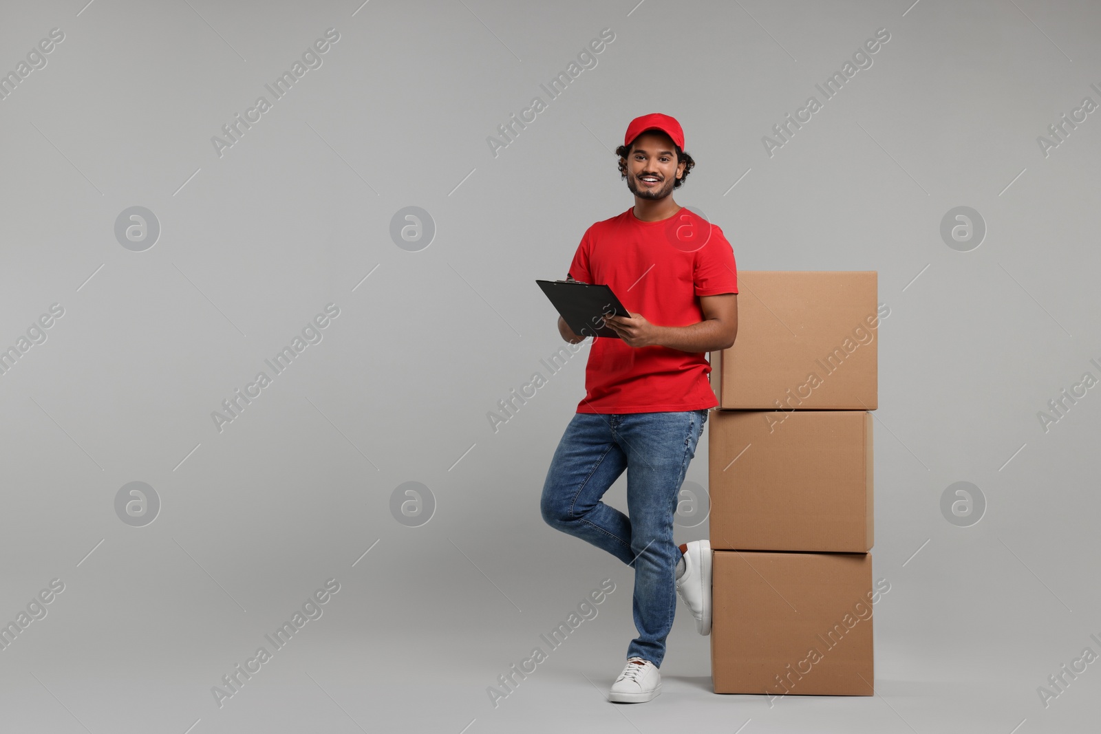 Photo of Happy courier with clipboard and stack of parcels on grey background, space for text