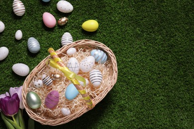 Photo of Easter basket with painted eggs and flowers on green grass, flat lay. Space for text