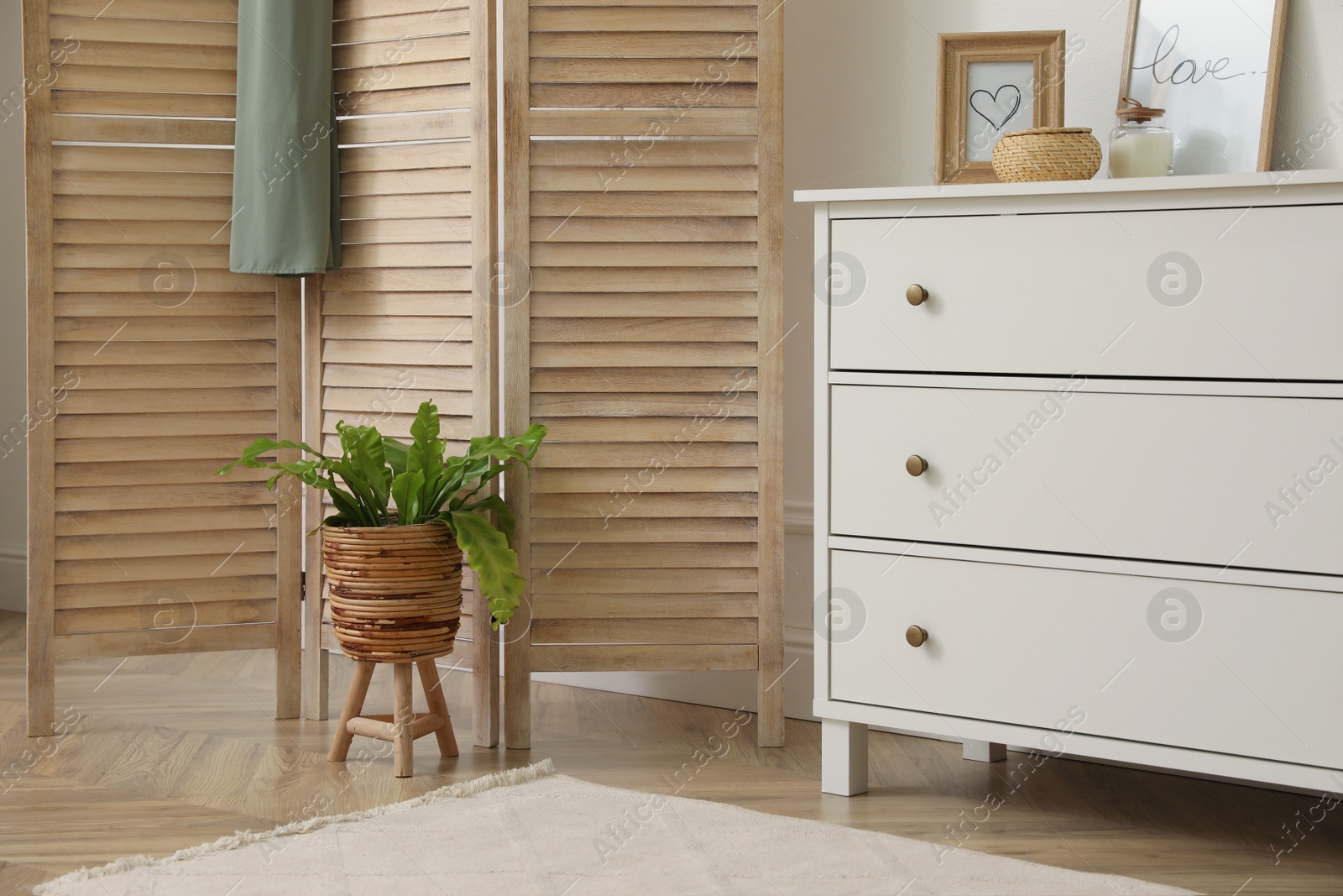 Photo of Stylish room with wooden folding screen and chest of drawers near white wall. Interior design