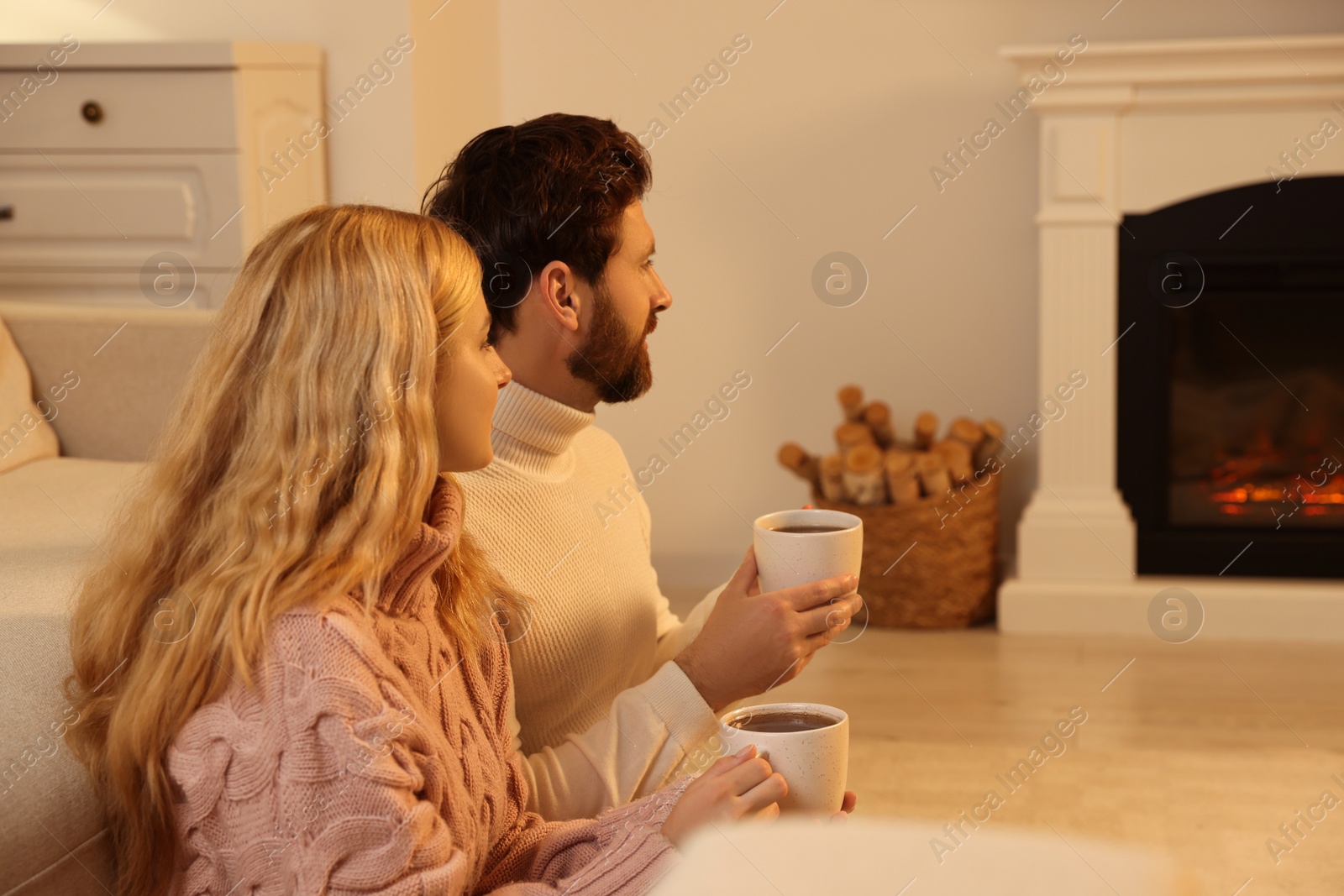 Photo of Lovely couple with hot drinks spending time together near fireplace at home