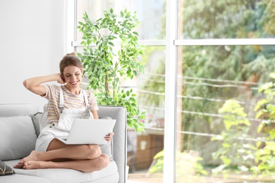Photo of Young woman with laptop sitting on sofa near window at home