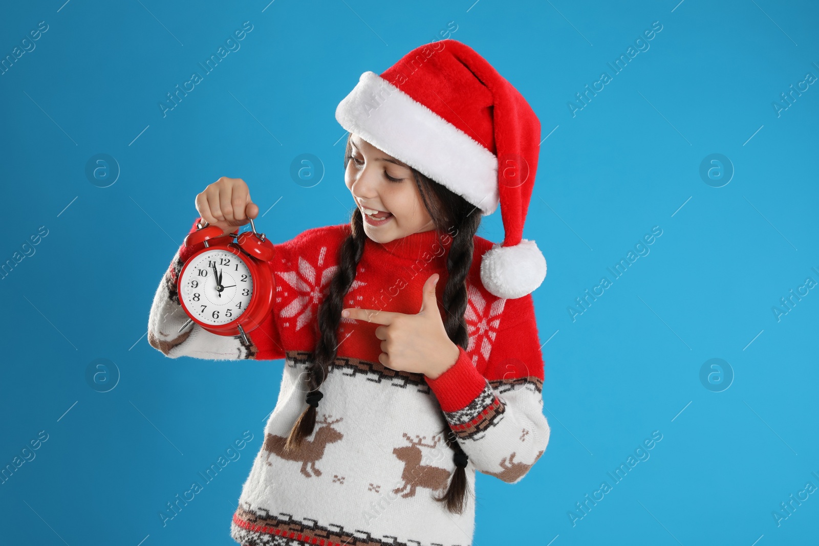 Photo of Girl in Santa hat with alarm clock on light blue background. New Year countdown