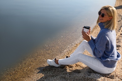 Happy woman in stylish sweater sitting on beach with cup of coffee