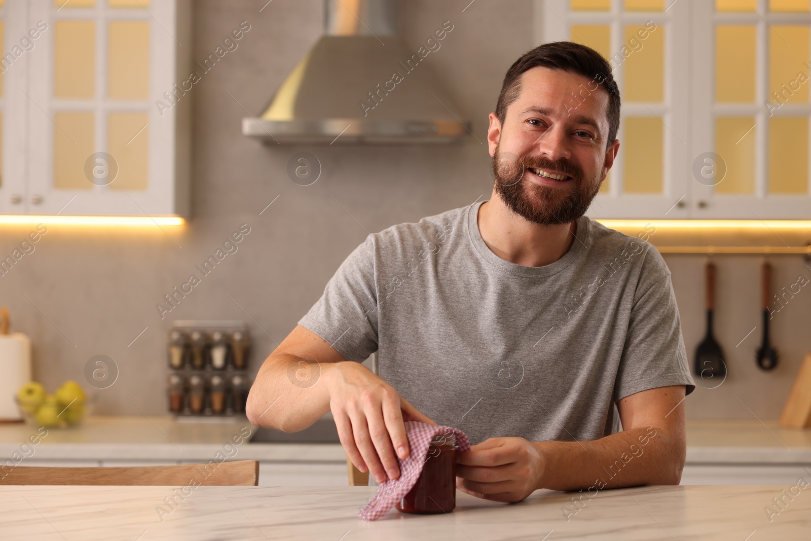 Photo of Man packing jar of jam into beeswax food wrap at table in kitchen