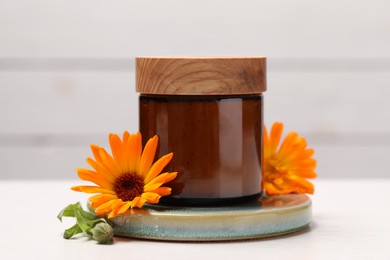 Jar of cosmetic product and beautiful calendula flowers on white table, closeup