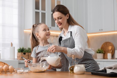 Photo of Making bread. Mother and her daughter pouring milk into bowl at wooden table in kitchen