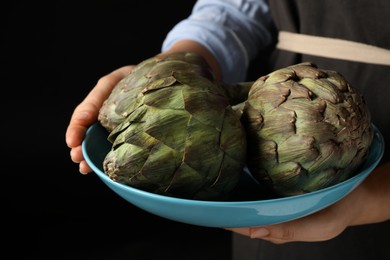 Woman holding bowl with fresh raw artichokes on black background, closeup