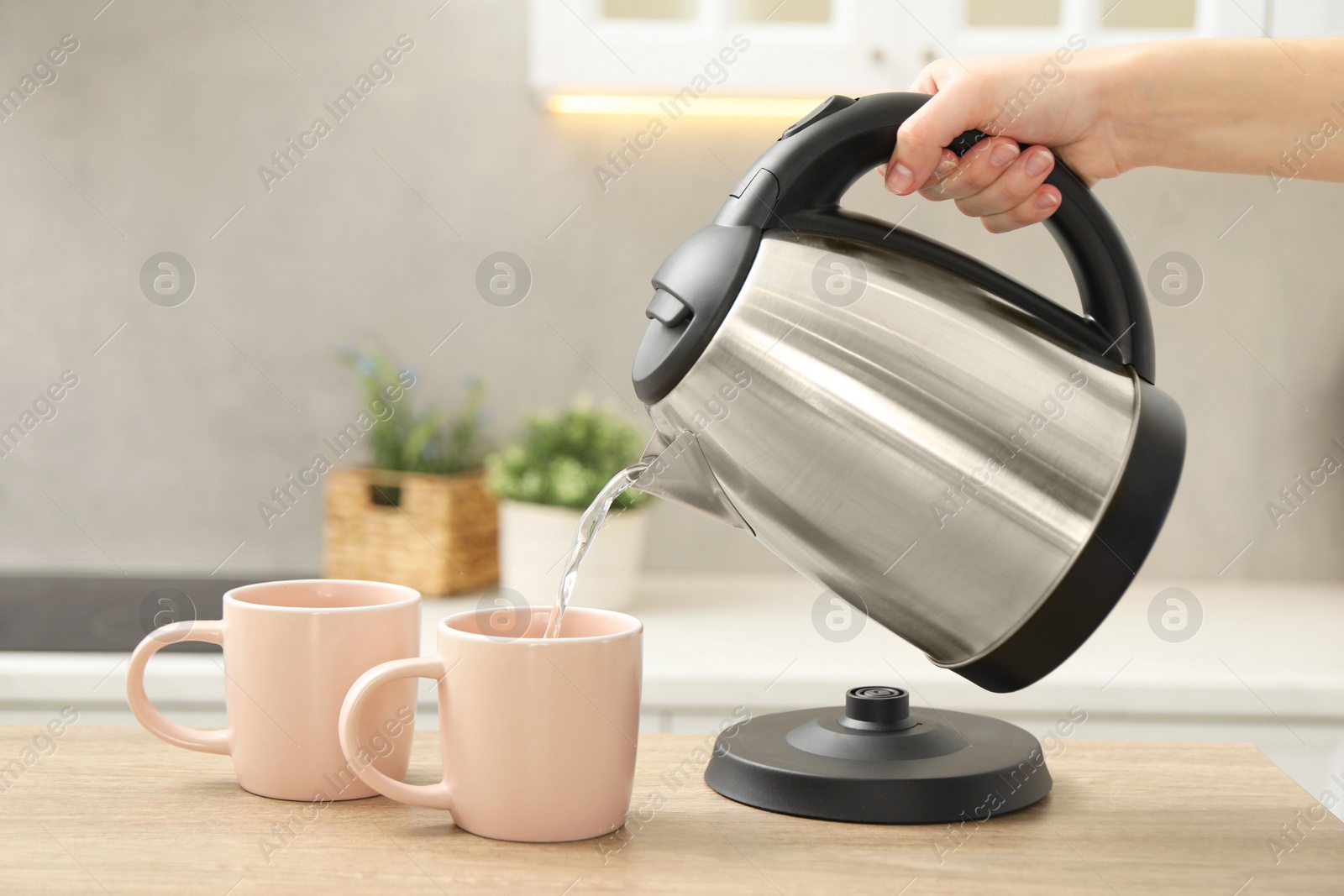 Photo of Woman pouring hot water from electric kettle into cup in kitchen, closeup