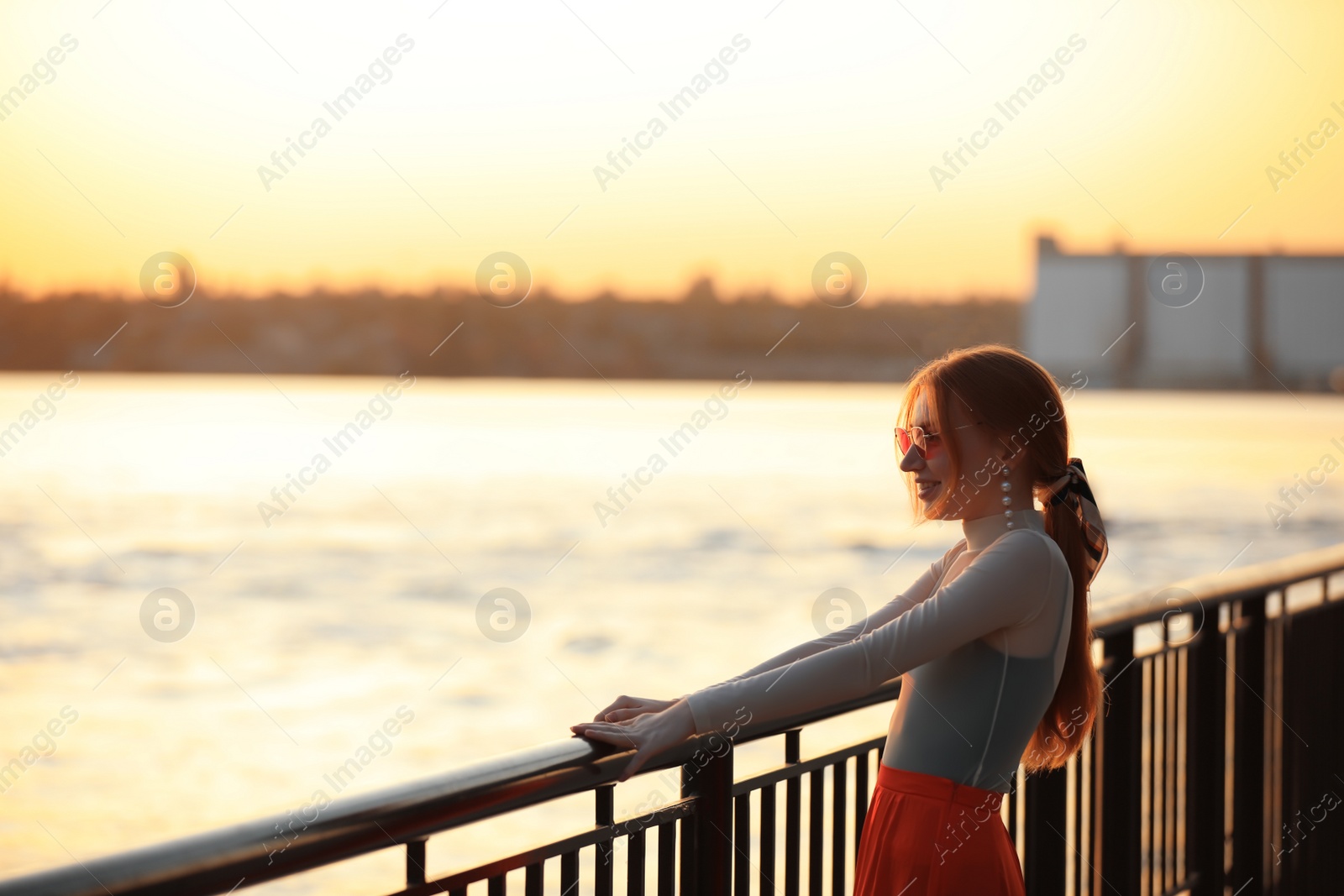 Photo of Young woman posing near railing on waterfront at sunset