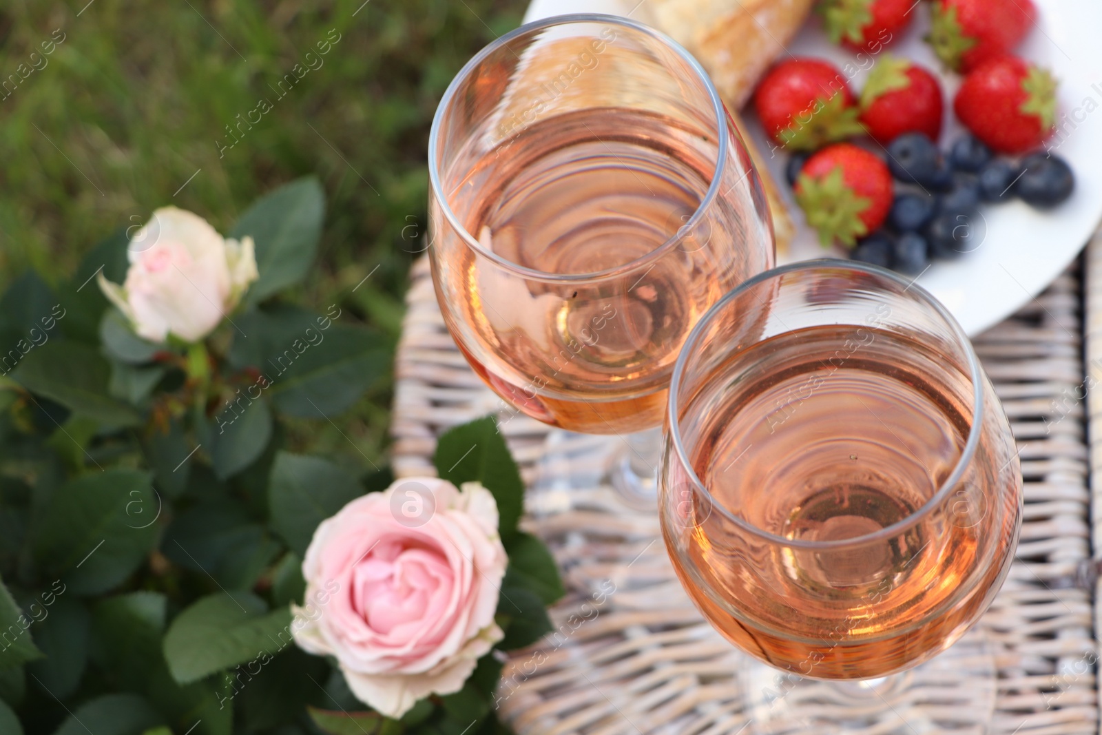 Photo of Flowers near glasses of delicious rose wine and food on picnic basket outdoors, closeup