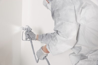 Decorator in protective overalls painting wall with spray gun indoors, closeup
