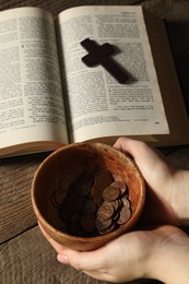 Photo of Donate and give concept. Woman holding bowl of coins, closeup. Bible and cross on wooden table