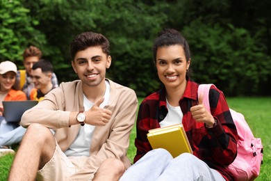 Happy young students showing thumbs up on green grass in park, selective focus